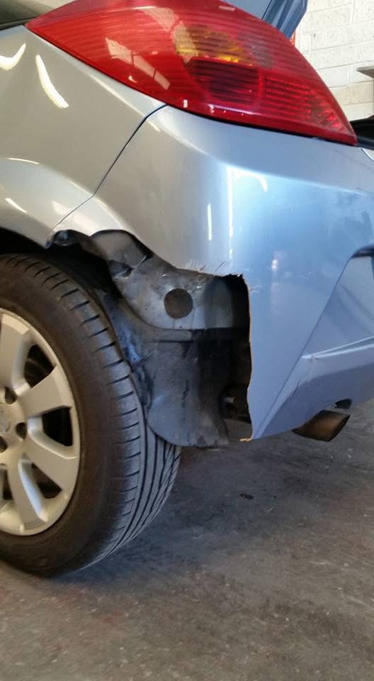 Before repair: damaged rear bumper with a missing section beside the rear wheel of a light blue modern classic